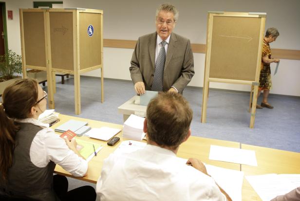 Ein Mann wirft seinen Stimmzettel in einer Wahlkabine ein, während Wahlhelfer anwesend sind.