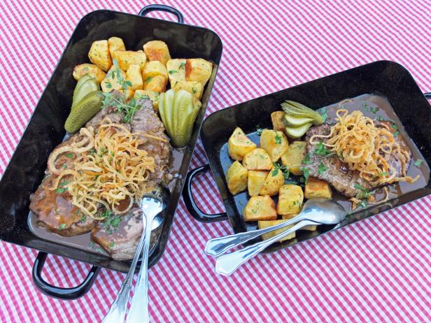 Zwei Bräter mit Zwiebelrostbraten, Bratkartoffeln und Gurken auf einer rot-weiß karierten Tischdecke.