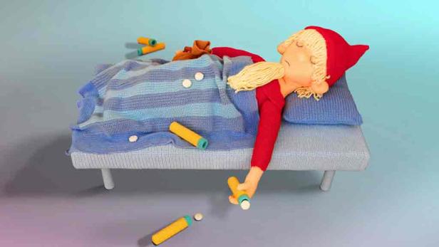 Eine Puppe mit roter Mütze liegt krank im Bett und hat Medikamente um sich herum.