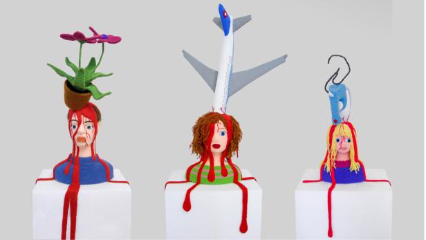 Drei gestrickte Puppenköpfe auf Sockeln, durchbohrt von einem Flugzeug, einer Blume und einem Bügeleisen.