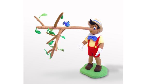 Eine gehäkelte Figur von Pinocchio mit langer Nase neben einem Ast mit einem blauen Vogel.
