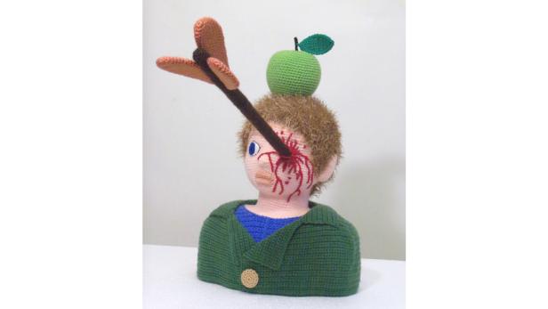 Eine gehäkelte Figur mit einem Apfel auf dem Kopf und einem Pfeil im Auge.