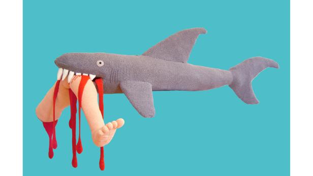 Ein gestrickter Hai hält ein blutiges Bein im Maul.