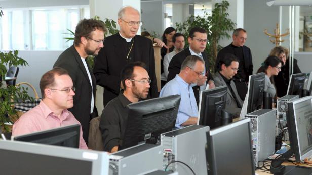 Eine Gruppe von Menschen, darunter ein Bischof, sitzt an Computern in einem Büro.