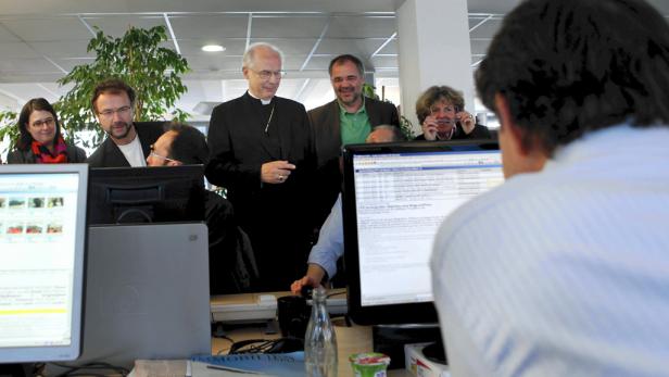 Eine Gruppe von Menschen steht um einen Schreibtisch mit Computern in einem Büro.
