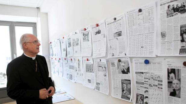 Ein Mann im Priestergewand steht vor einer Wand mit Zeitungsartikeln.