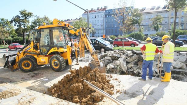Ein gelber Bagger hebt Erde aus einer Baugrube, während Bauarbeiter die Arbeiten beobachten.