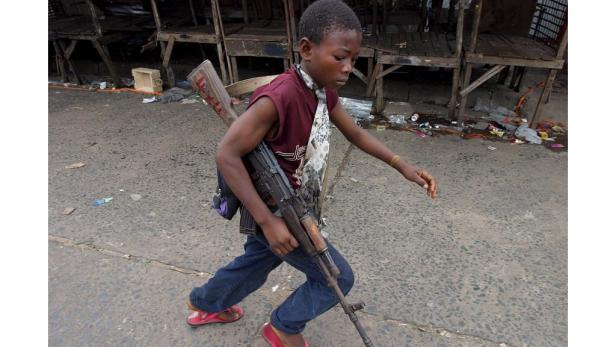 Ein junger Mann trägt ein Gewehr und eine Bazooka über der Schulter.