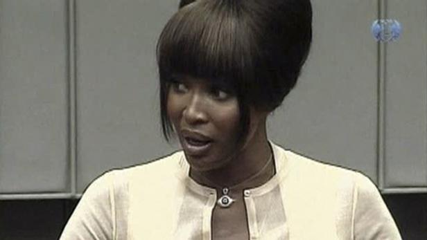 Naomi Campbell mit auffälliger Frisur und hellem Oberteil vor grauem Hintergrund.