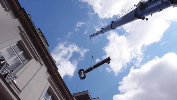 Ein Kran hievt einen riesigen, alten Schlüssel vor blauem Himmel in die Höhe.