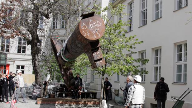 Eine große, rostige Skulptur mit arabischer Schrift steht im Freien vor einem Gebäude.