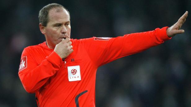 Ein Fußballschiedsrichter in roter Uniform pfeift und hebt den Arm.