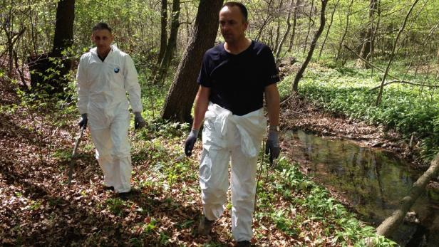 Zwei Personen in Schutzanzügen gehen durch einen Wald entlang eines kleinen Baches.