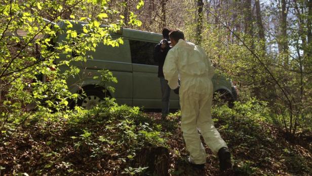 Ein Mann in Schutzanzug untersucht einen Van in einem Waldstück.