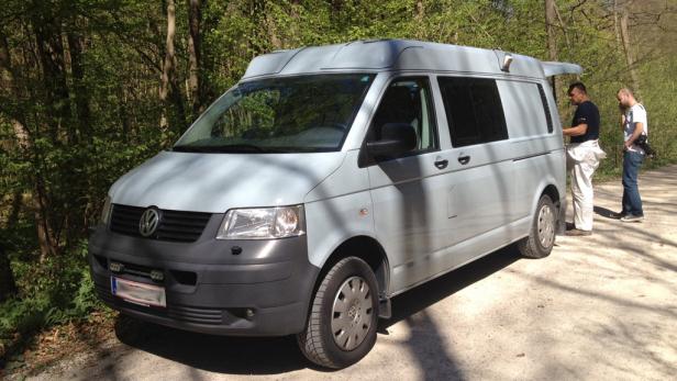 Ein grauer VW-Bus steht am Waldrand, zwei Männer stehen daneben.