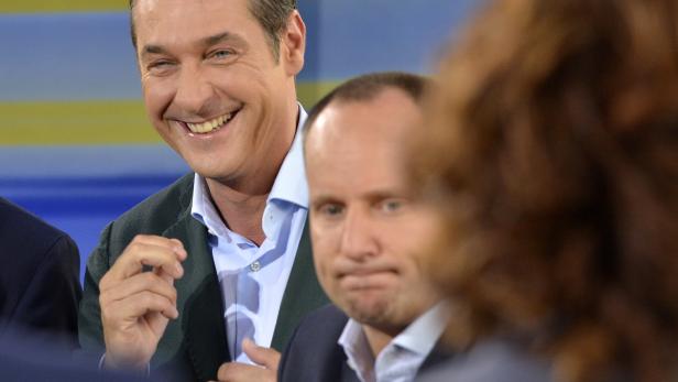 Zwei Männer in Anzügen, einer lächelt, der andere blickt ernst.