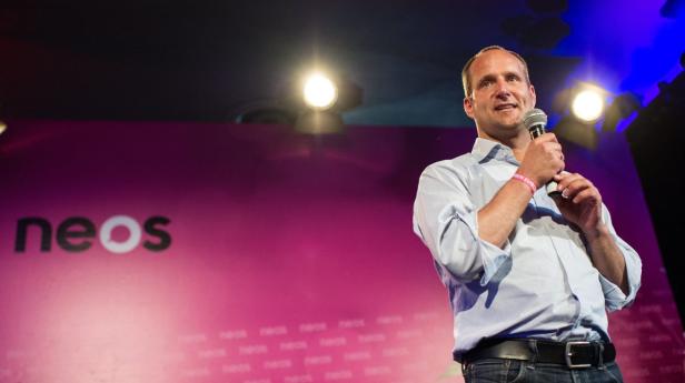 Ein Mann spricht auf einer Bühne vor einem pinkfarbenen Hintergrund mit dem Neos-Logo in ein Mikrofon.