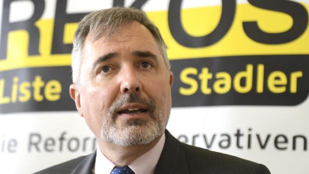 Ein Mann mit Bart steht vor einem Banner mit der Aufschrift „REKOS Liste Stadler“.