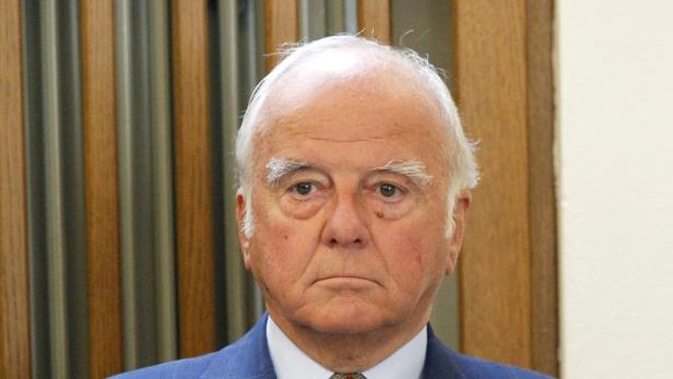 Ein älterer Mann mit weißem Haar und einem blauen Anzug blickt in die Kamera.