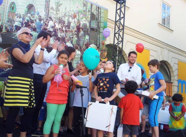 Eine Gruppe von Kindern steht mit Luftballons auf einer Bühne, während Erwachsene applaudieren.