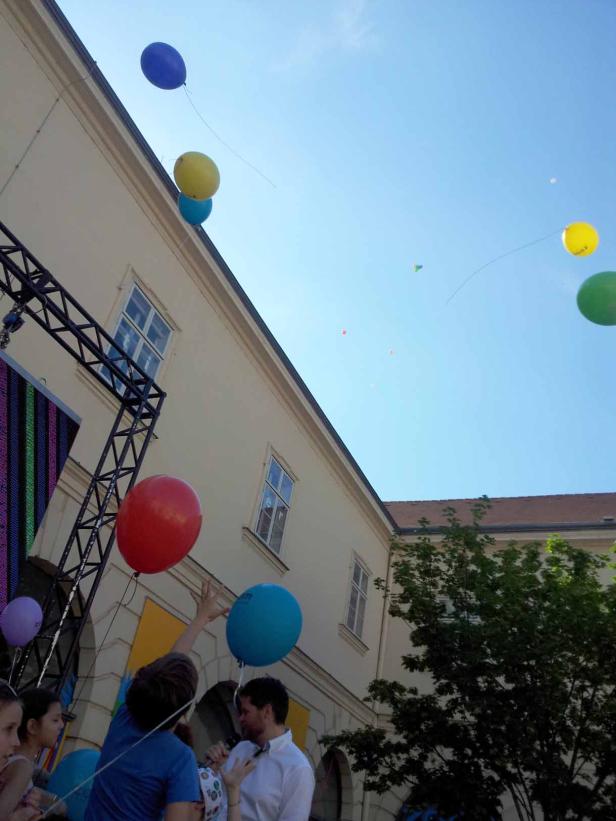 Menschen lassen bunte Ballons vor einem Gebäude in den Himmel steigen.