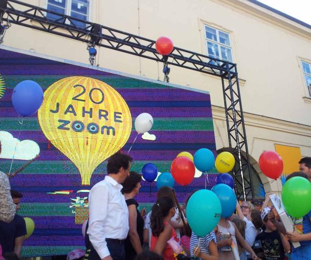 Eine Menschenmenge versammelt sich vor einer Bühne mit der Aufschrift „20 Jahre Zoom“ und vielen bunten Ballons.