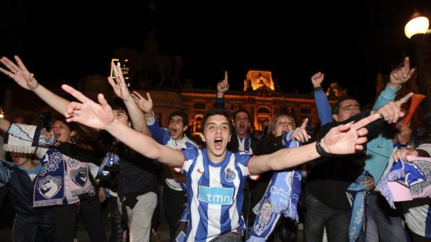 Eine Gruppe jubelnder Fußballfans feiert nachts mit Schals und Trikots vor einem Gebäude.