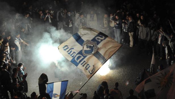 Eine Menschenmenge jubelt inmitten von Rauch und schwenkt eine blau-weiße Flagge.