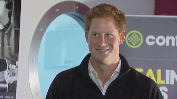 Prinz Harry lächelt vor einem runden Fenster und einem Logo.