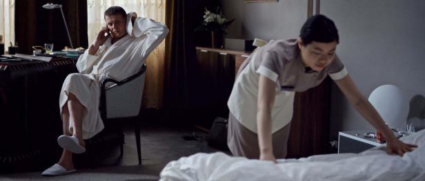 Ein Mann im Bademantel telefoniert, während ein Zimmermädchen das Bett macht.