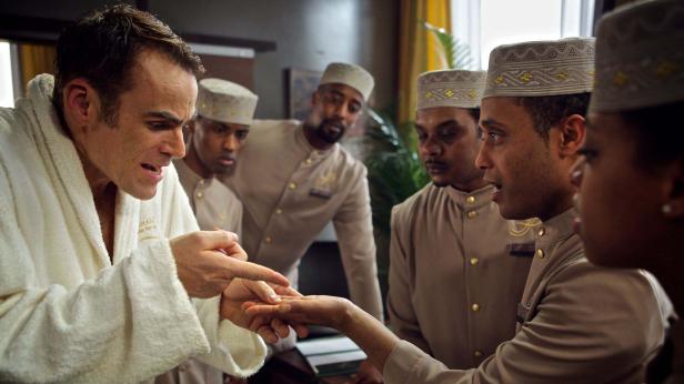 Ein Mann im Bademantel gestikuliert vor einer Gruppe Hotelangestellter in Uniformen.