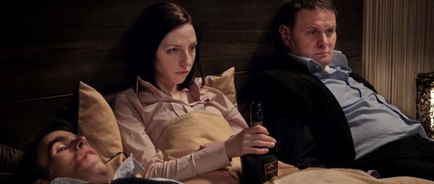 Eine Frau hält eine Flasche, während zwei Männer im Hintergrund liegen.