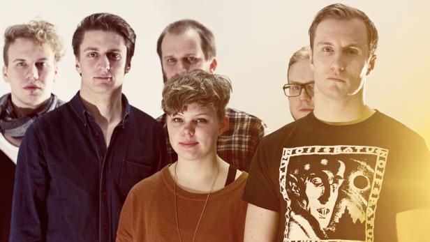 Eine Gruppe von sechs jungen Erwachsenen posiert für ein Gruppenfoto vor einem hellen Hintergrund.