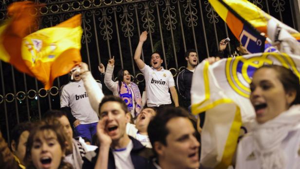 Eine Gruppe jubelnder Real Madrid-Fans mit Trikots und Fahnen vor einem Gitter.