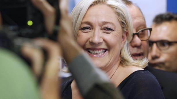 Marine Le Pen lächelt in die Kamera, umgeben von Fotografen und anderen Personen.