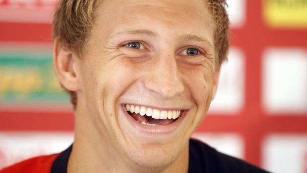 Ein lachender Mann mit blonden Haaren und blauen Augen vor einem roten Hintergrund.