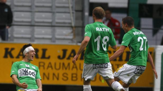 Ein jubelnder Fußballspieler mit Kopfverband wird von seinen Mannschaftskollegen Rath und Röche gefeiert.