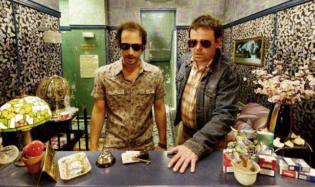 Zwei Männer mit Sonnenbrillen stehen an einem Rezeptionstresen in einem Hotel.