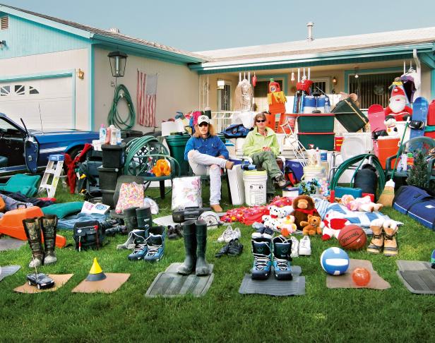 Zwei Personen sitzen vor einem Haus, umgeben von Gegenständen für einen Garagenflohmarkt.