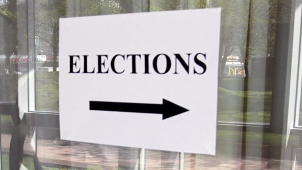 Ein Schild mit der Aufschrift „Elections“ und einem Pfeil nach rechts hängt an einer Glastür.