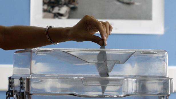 Eine Hand wirft einen Stimmzettel in eine durchsichtige Wahlurne.