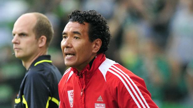 Ein Fußballtrainer in roter Jacke gestikuliert neben einem Schiedsrichter.