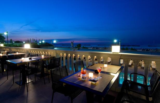 Ein abendlicher Blick von einer Terrasse auf das Meer mit beleuchteten Tischen und Cocktails.