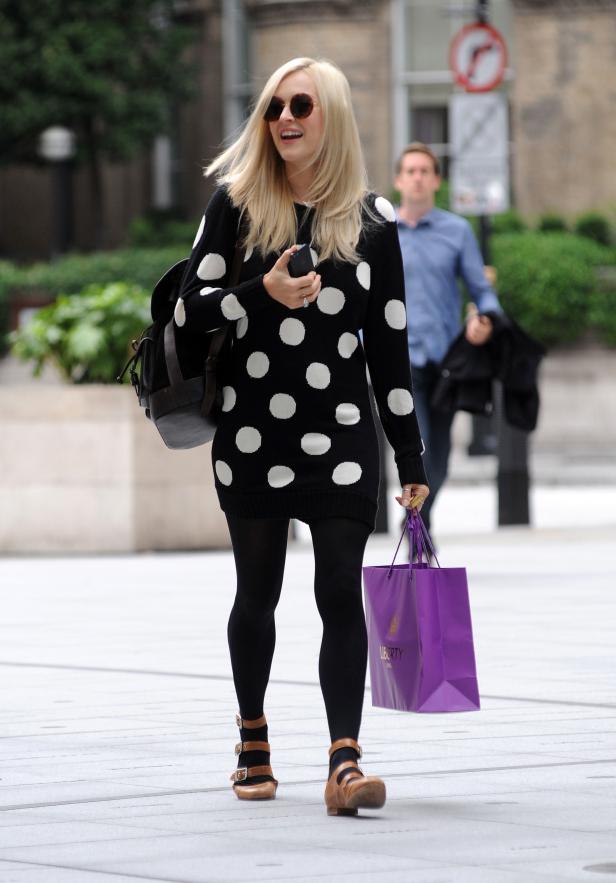 Eine Frau mit Sonnenbrille, einem gepunkteten Kleid und einer Einkaufstasche von Liberty läuft auf der Straße.