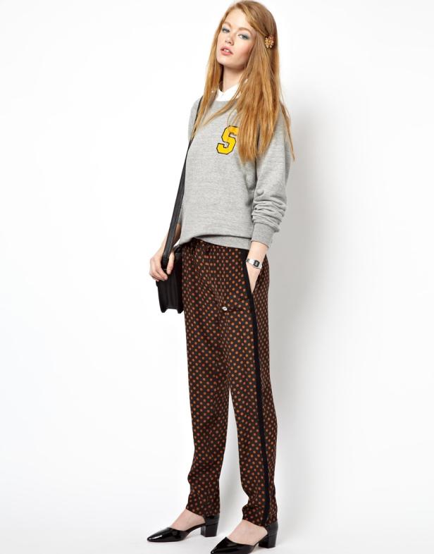 Eine junge Frau trägt einen grauen Pullover mit einem gelben „S“ und eine braun gepunktete Hose.