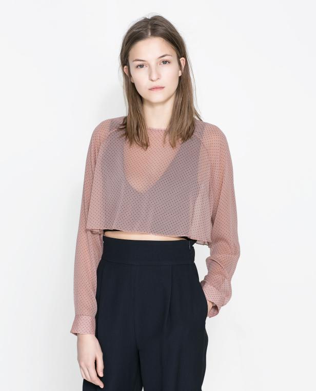 Eine Frau posiert in einer rosafarbenen, gepunkteten Bluse und einer schwarzen Hose vor einem weißen Hintergrund.