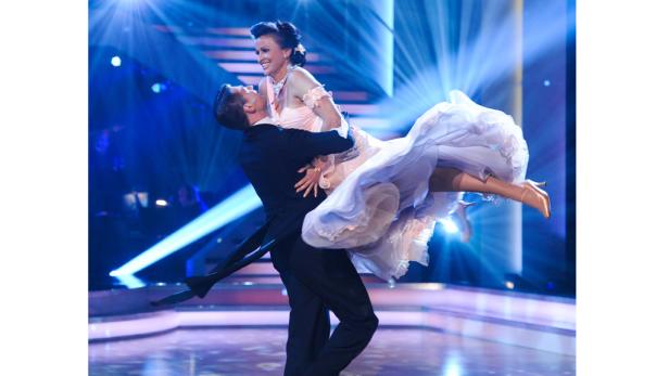 Ein Mann im Anzug hebt eine Frau im Ballkleid auf einer Bühne hoch.