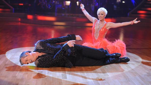 Ein Mann liegt erschöpft auf dem Tanzparkett, während eine Frau im roten Kleid jubelt.