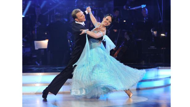 Ein Tanzpaar führt eine elegante Darbietung auf einer Bühne mit Orchester im Hintergrund auf.