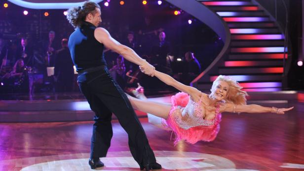 Ein Tanzpaar führt eine Hebefigur auf einer Bühne auf, begleitet von einem Orchester.